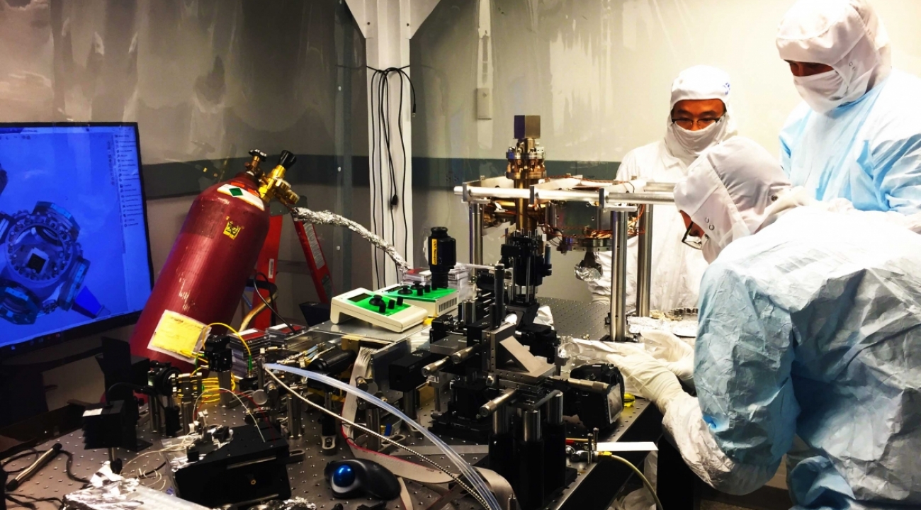 Marko Centina, Kai Hudek, and Daiwei Zhu assembling an ion trap vacuum system in a clean room. Their suits prevent contaminants from entering the apparatus.