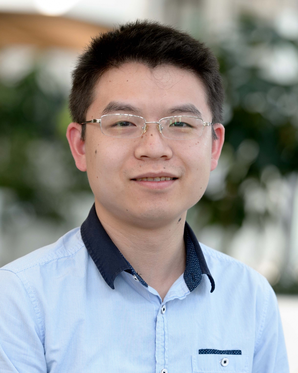 A man in a blue shirt stands in front of an out-of-focus outdoor background.