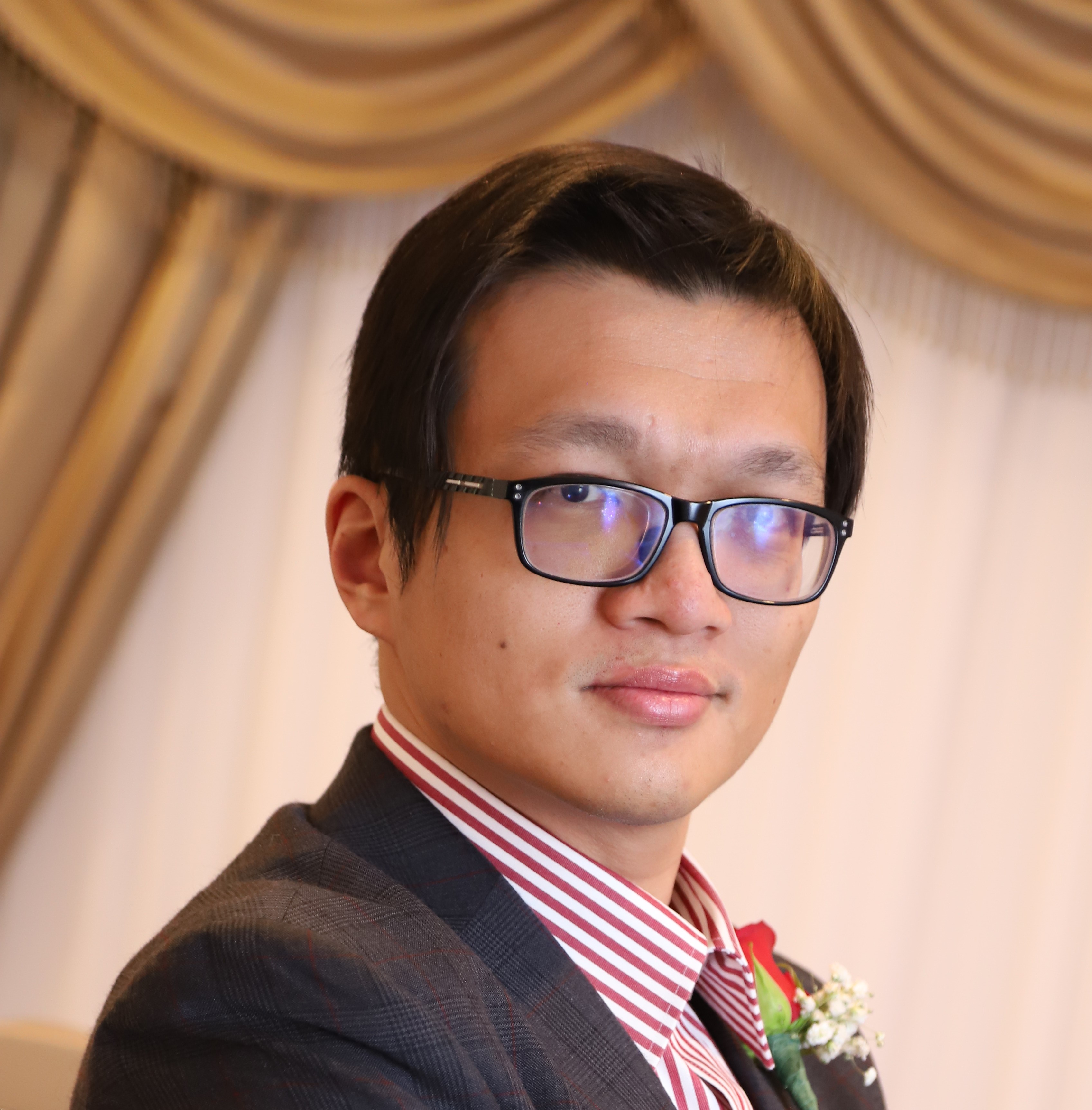 A young man sits posed in front of draped golden and white fabric. He wears glasses, a red and white striped shirt, a wool suit jacket, and a rose bouttaniere.  He is faintly smiling.