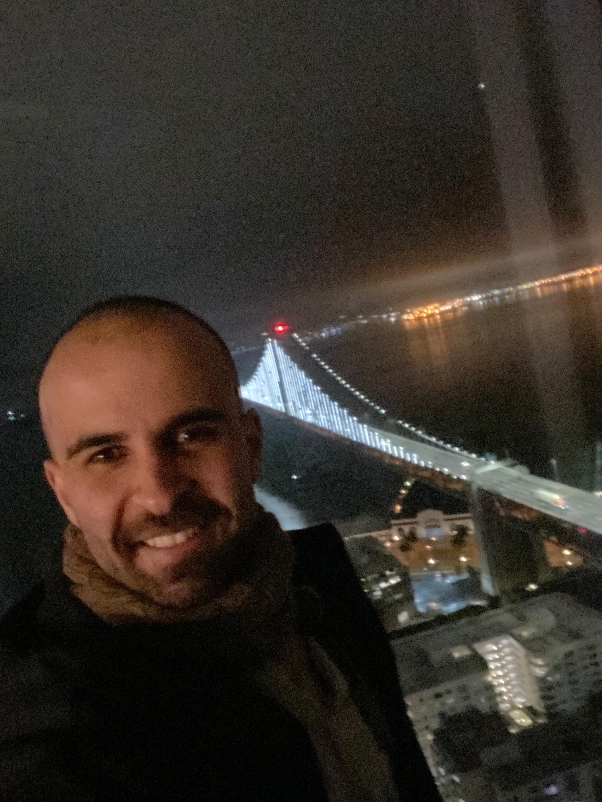 A smiling man appears with a bridge in the background, night time setting.