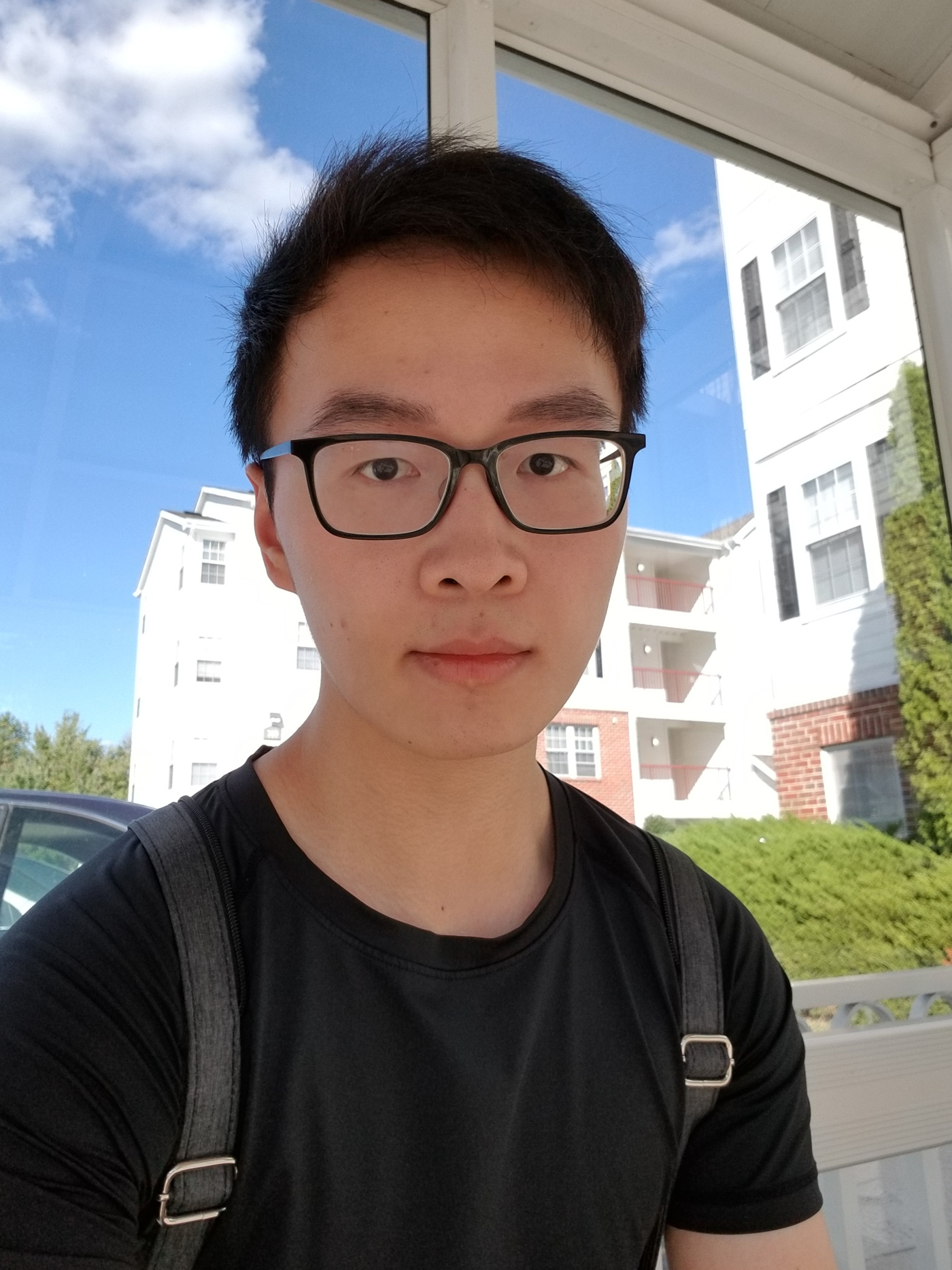 A photo of Kaixin Huang, who looks intently at the camera. He wears a black t-shirt and backpack. The sky is blue and cloudy behind him.