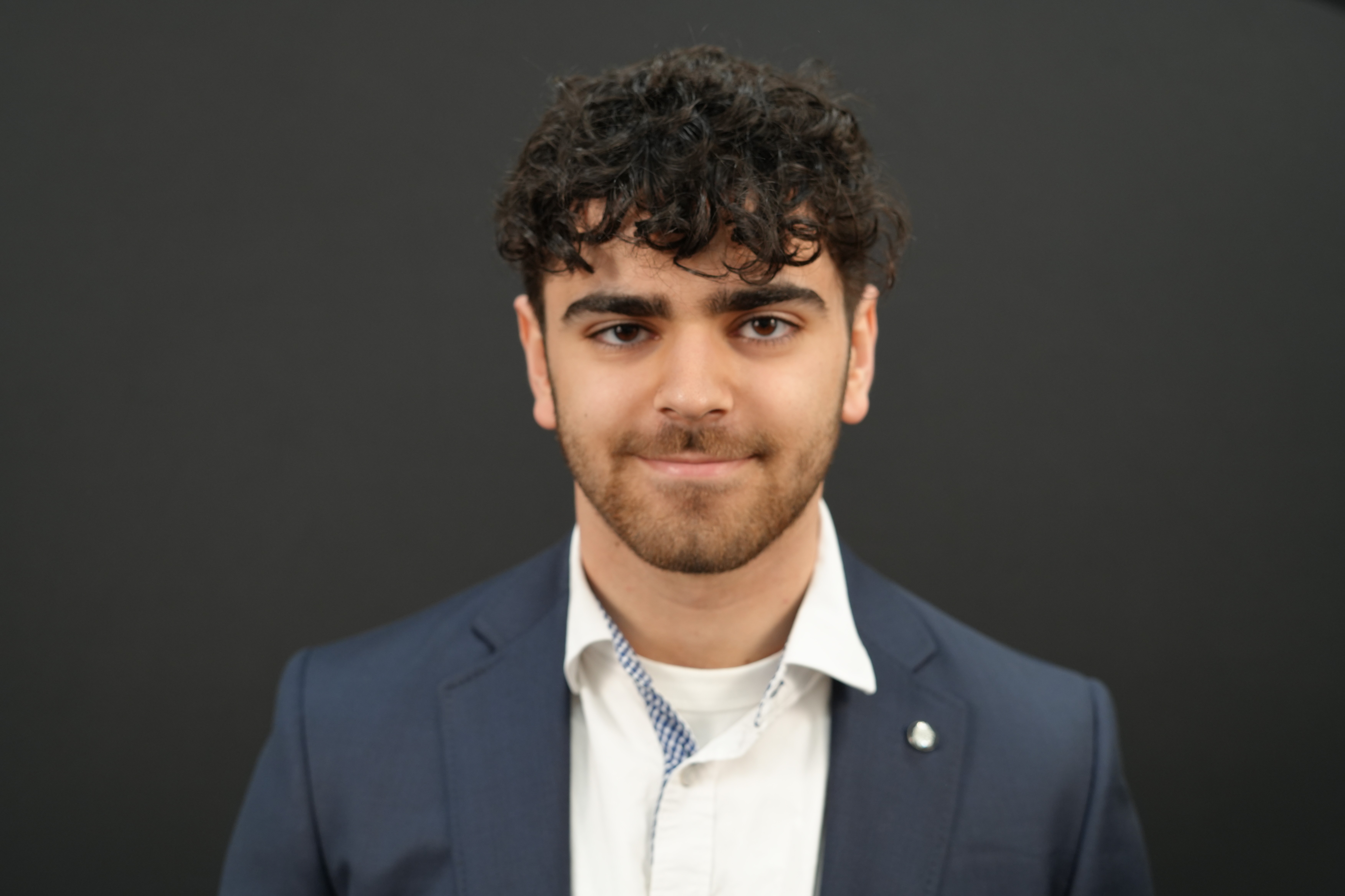 A photo of Valery Simonyan, wearing a casual navy blue suit jacket.  His hair is dark and curly, he has a light beard, and he is slightly smiling.  The background is grey. 