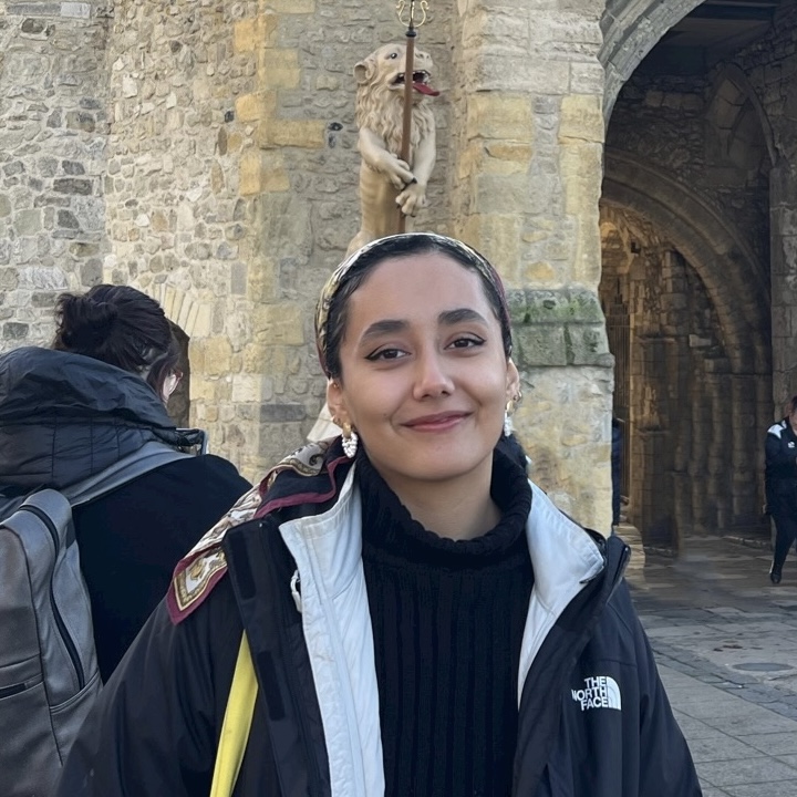 A smiling young woman appears standing in front of a stone wall.