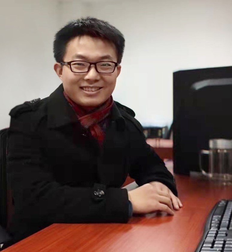 A smiling young Asian man at a desk with short dark hair and glasses.