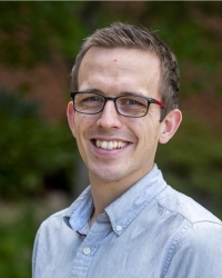 Head and shoulder of a smiling, light skinned man wearing glasses in an outdoors setting