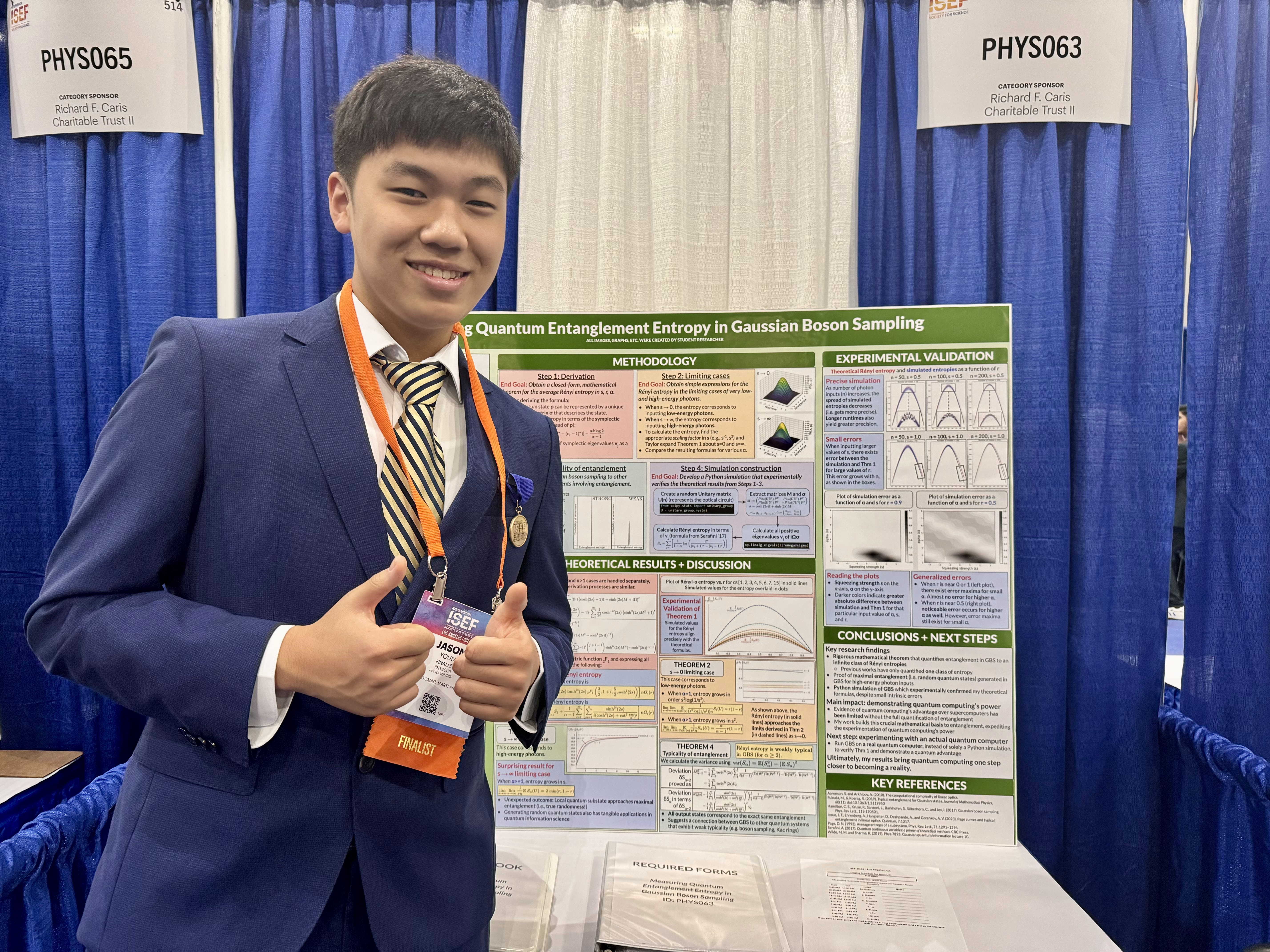 A young man in a blue blazer stands in front of a poster showing graphs and information while doing a thumbs up with both hands.