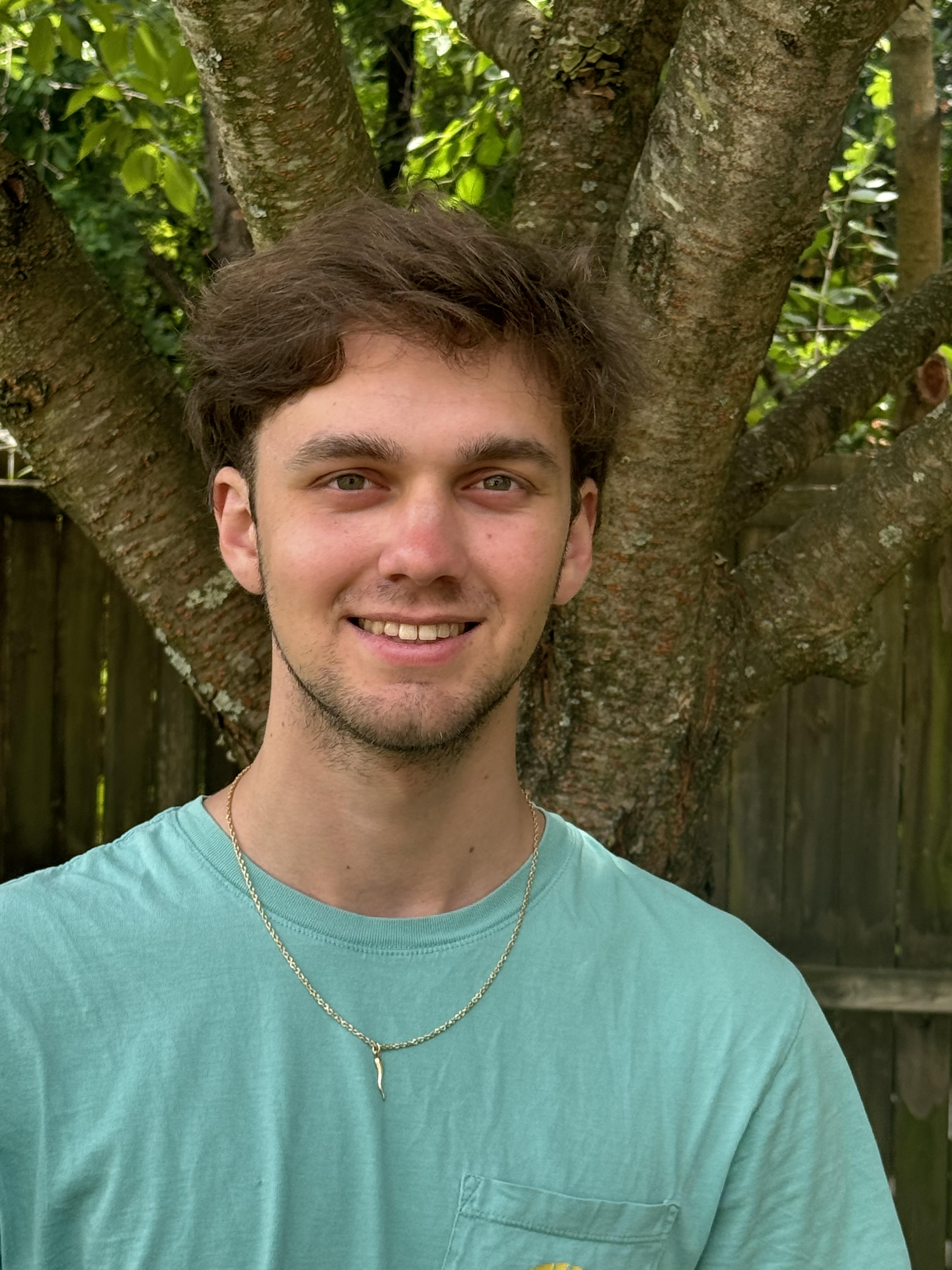 A smiling young man with light wearing a green teeshirt is pictured outdoors, with a tree in the background.