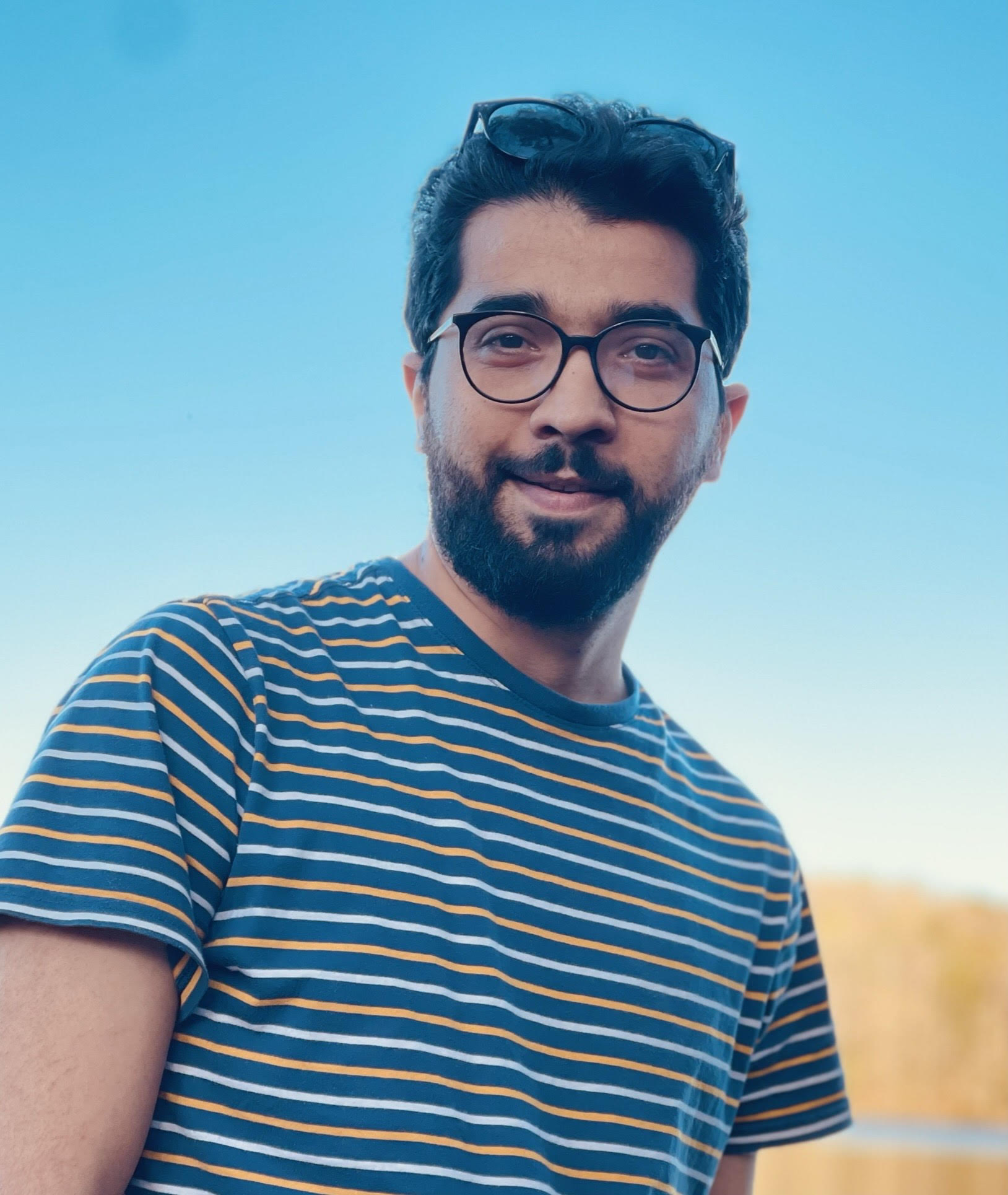 A smiling, bearded young man wearing glasses and a striped shirt appears outdoors with a blue sky background.