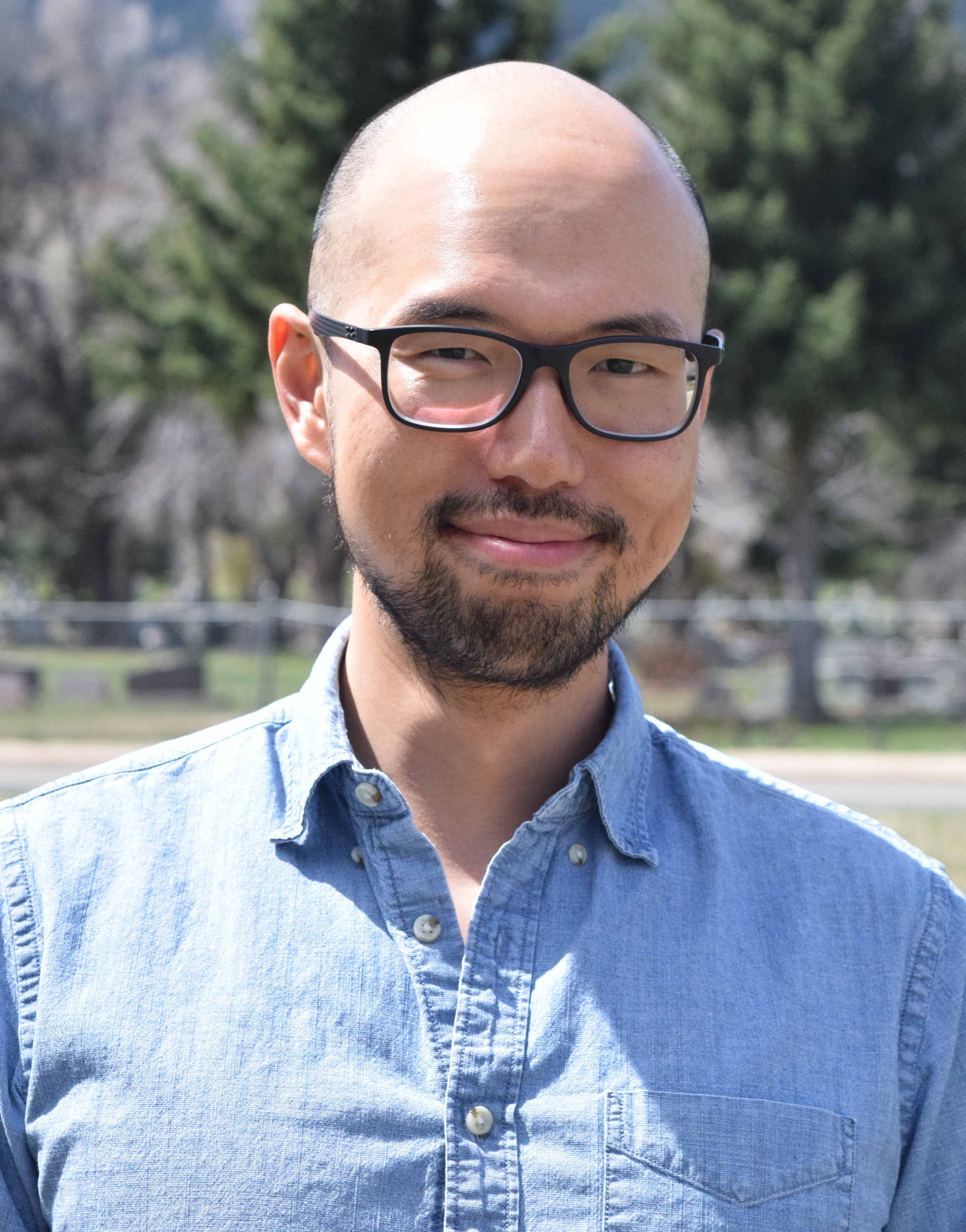 A man in glasses and a blue denim shirt stands in front of a fence and trees.