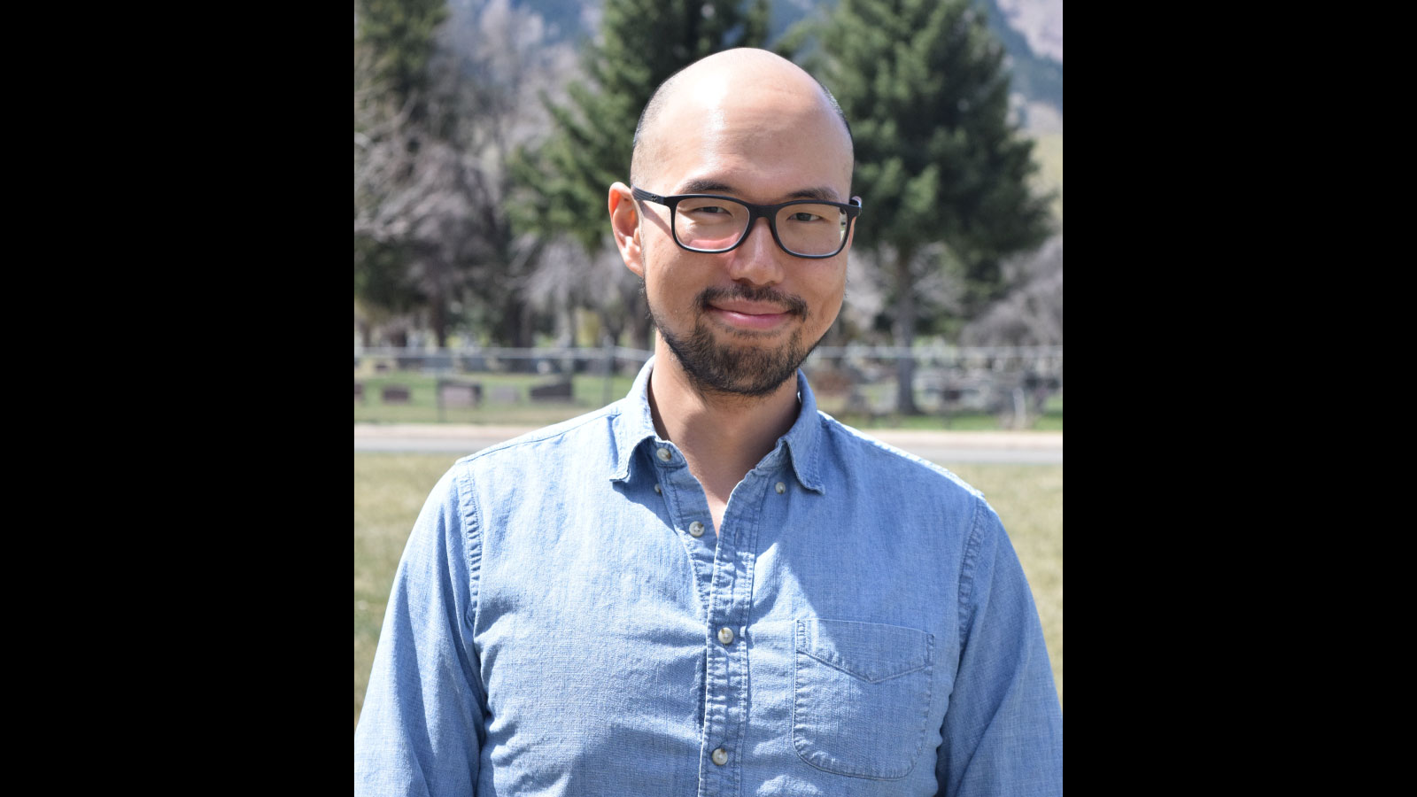 A man in glasses and a blue denim shirt stands in front of a fence and trees.