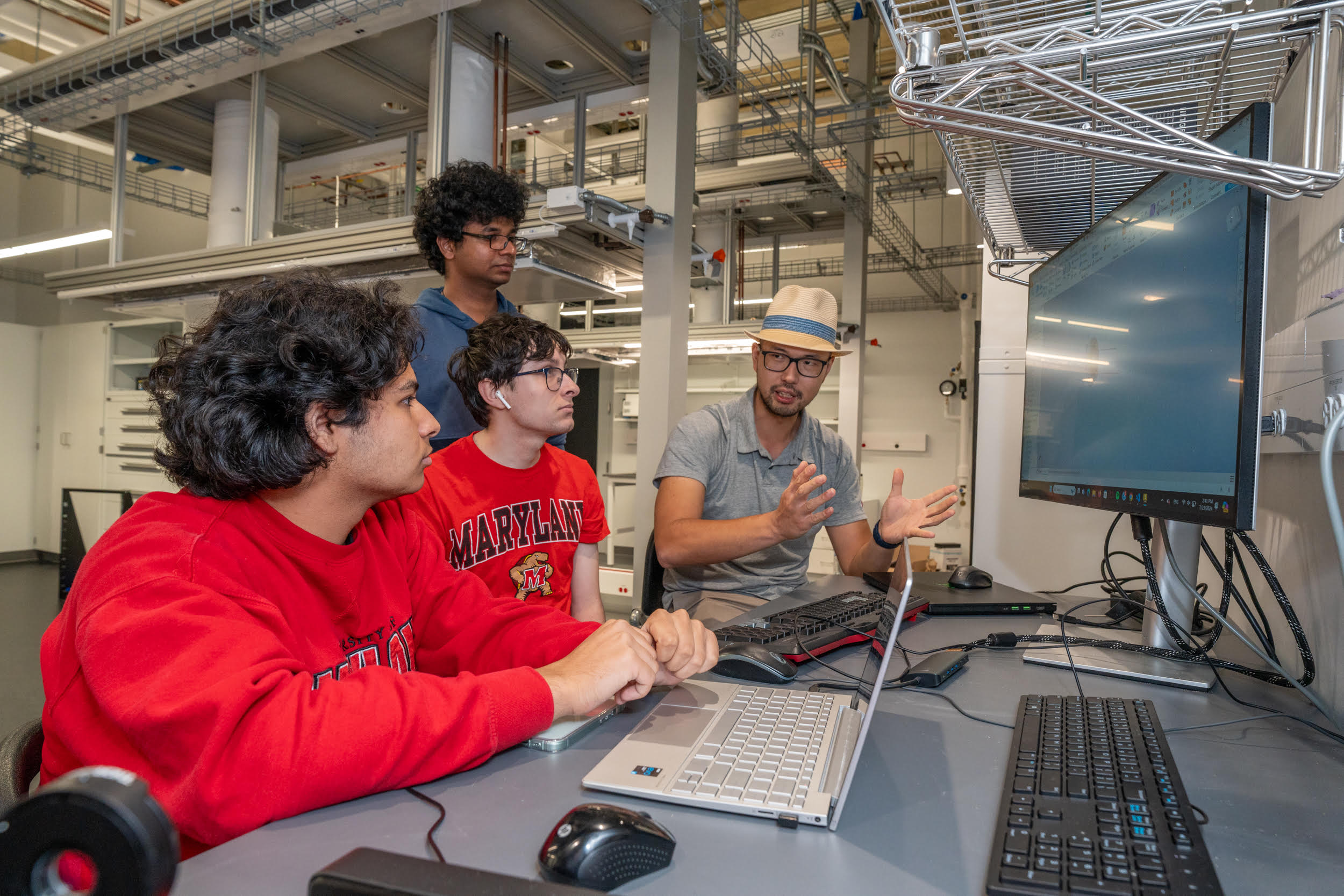 A man in a hat talks to three students as they set in front of computers with clean laboratory space behind them.