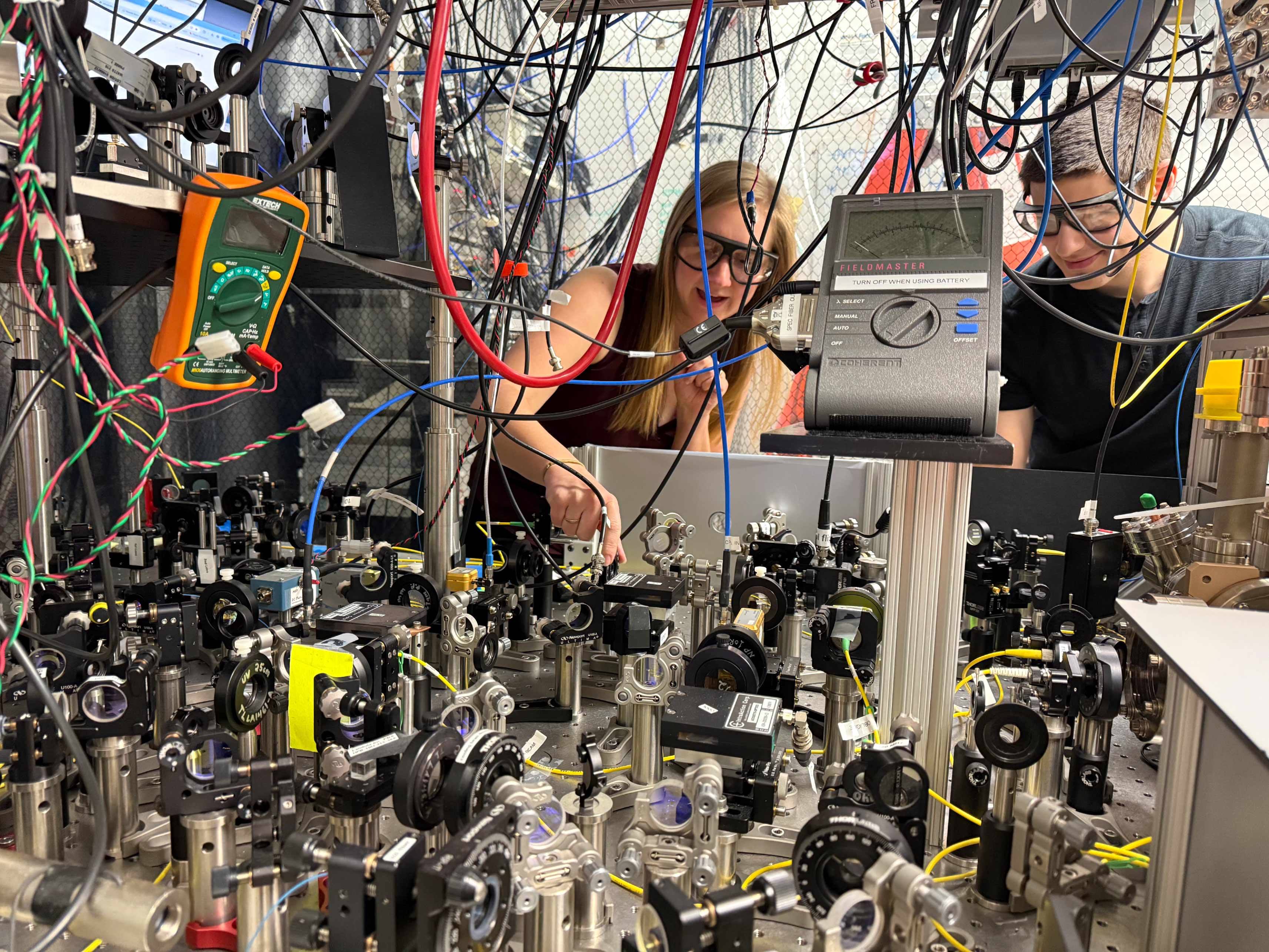 A woman and a man wearing googles are partially obscured behind dangling cables and a table full of scientific equipment and optical devices, such as lenses and mirrors.