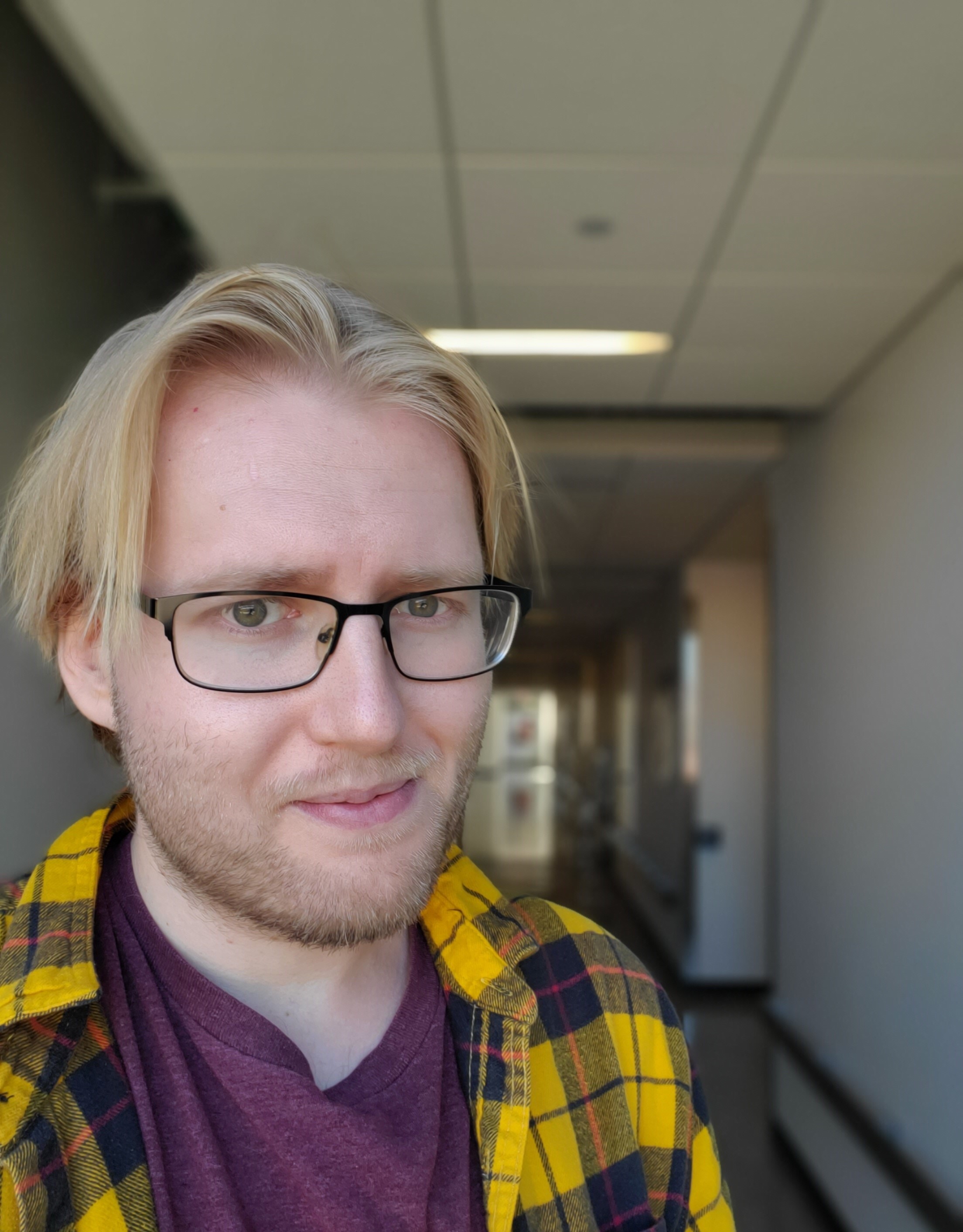 A young man with blond hair and glasses, in a hallway, smiles at the camera.