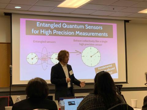 Bringewatt in a white shirt and blazer stands in front of a projector screen showing a slide that says "Entangled Quantum Sensors for High Precision Measurements" and shows basic graphics.