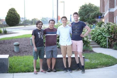 Four men stand on a sidewalk in front of a bench with their arms around each others sholders.