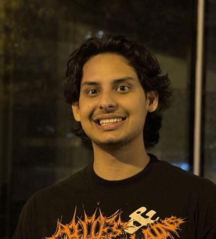 A smiling young man with brown skin and dark hair is pictured in an indoor setting