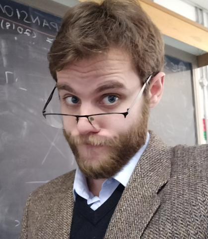 A bearded young man with light brown hair peers over his glasses, with a questioning look, and a blackboard in the background.